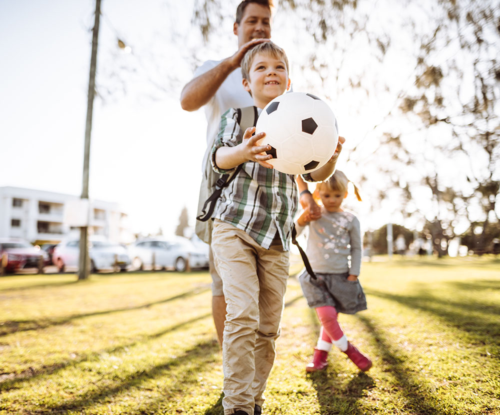 A father walks through a park with his young kids and a soccer ball